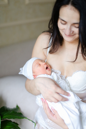 Young smiling mother wearing white clothes holding newborn baby. Concept of motherhood and happiness.の写真素材