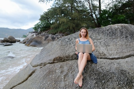 Young businesswoman working with laptop on empty beach and sitting on stone. Concept of resting on morning sea and summer vacations, modern technology.の写真素材