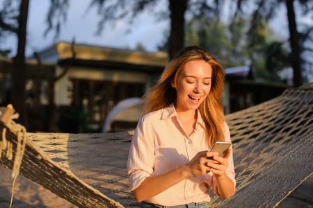 Young happy girl using smartphone and sitting in wicker hammock on tropical resort. Concept of modern technology and summer vacations in Thailand.の写真素材