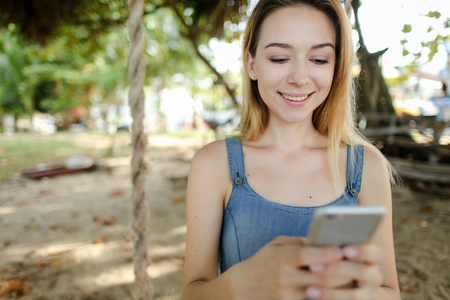 Young american female person riding swing and using smatrphone, sand and tree in background. Concept of summer vacations and modern technology.の写真素材