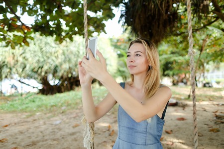 Young beautiful woman riding swing and making selfie by smartphone, sand and trees in background. Concept of summer vacations and modern technlogy, leisure time.の写真素材