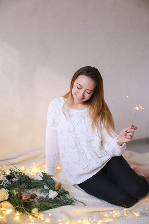 Young pretty woman sitting with bengal light near twinkling yellow garlands and green wreath. Concept of handmade decorations for Christmas and New Year holidays.の写真素材