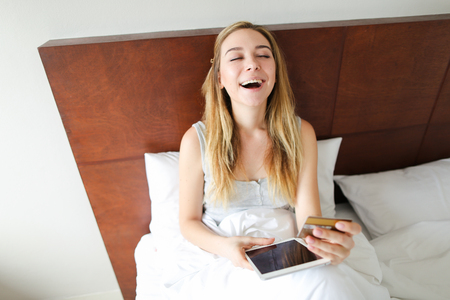 Young female person going shopping online with tablet and debit card, sitting in bed. Concept of purchasing on internet and webstore.の写真素材