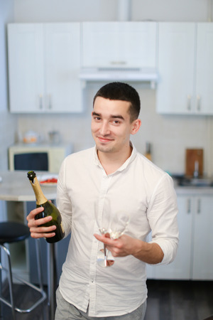 Young man standing in kitchen with champagne and wineglass, wearing white shirt. Concept of celebrating birthday, alcohol and youth.の写真素材