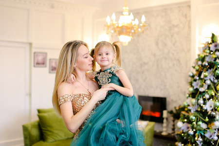 Young woman kissing little daughter, wearing dress and standing near Christmas tree. Concept of celebrating winter holidays, New Year and fashionable look.の写真素材