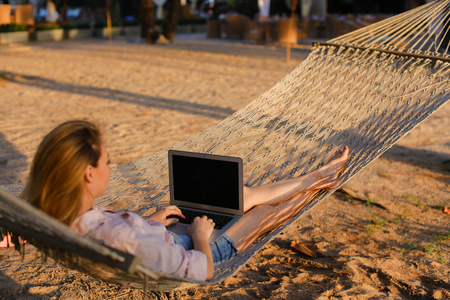 Young caucasian woman lying in white wicker hammock with sand background and using laptop. Concept of modern technology and summer vacations, resting on beach.の写真素材