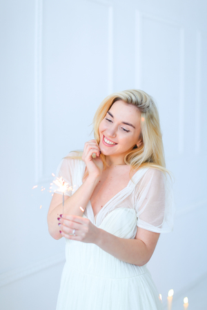 Blonde smiling woman keeping bengal light and wearing white dress. Concept of happiness and celebrating holidays.の写真素材