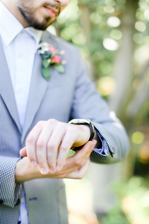 Caucasian groom with beard looking at watch and wearing grey suit. Concept of wedding day and time.の写真素材