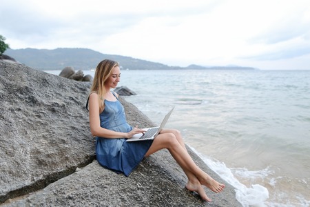 Young woman using laptop and sitting on stone near sea, empty morning beach. Concept of modern technology and working on nature, summer vacations.の写真素材