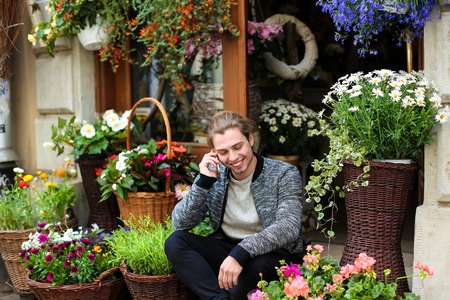 Young caucasian man speaking by smartphone and sitting at flower shop near bouquets in baskets. Concept of business and modern technology.の写真素材