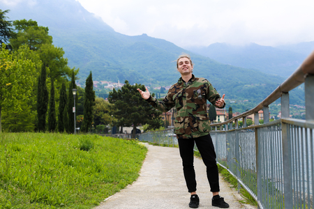 Young caucasian man wearing camouflage jacket standing near banister, lake Como and Alps mountain in background. Concept of tourism and European landscapes.の写真素材
