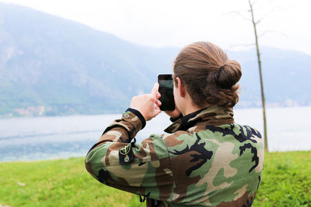 Young male tourist wearing camouflage jacket, taking photo by smartphone, lake Como and Alps mountain in background. Concept of modern technology, tourism and nature.の写真素材