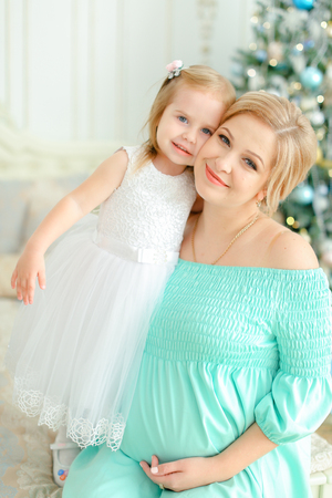 Pregnant mother wearing blue dress and hugging little daughter near Christmas tree. Concept of winter holidays and seasonal photo session.の写真素材