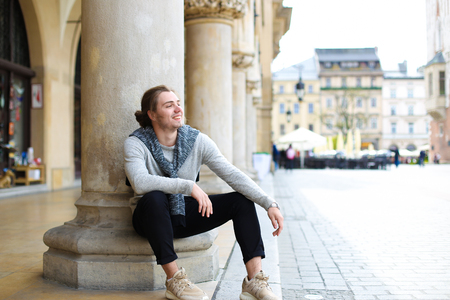 Young caucasian male tourist wearing casual clothes and sitting near columns on Main Square in Krakow, Poland. Concept of urban lifestyle, tourism and european youth.の写真素材