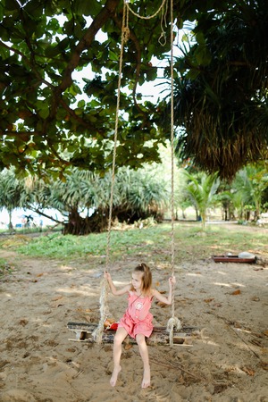 Female child riding on swing on sand background, wearing pink dress. Concept of childhood and summer vacations on exotic resort.の写真素材