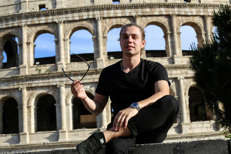 Young male tourist sitting near Colosseum background in Rome, Italy. Concept of cheapest tours to Europe and traveling to ancient historic landmarks.の写真素材
