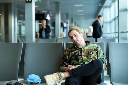 Caucasian man sitting in waiting room at airport and listening to music with white earphones, wearing camouflage shirt. Concept of traveling and cheap tickets, law cost flights.の写真素材