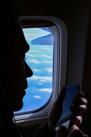 Closeup silhouette of young man using smartphone near window in airplane. Concept of modern...