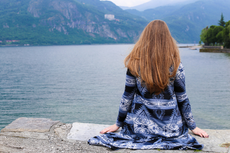 Back view of young woman with long hair sitting near lake Como, mountain in background. Concept of summer vacations.の写真素材