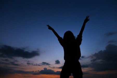 Silhouette of young woman in sunset dark blue sky with clouds background. Concept of freedom and calm evening.の写真素材