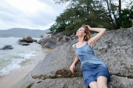 Young happy woman lying on stones on sea shore in morning, wearing jeans sundress. Concetp of relaxing on nature and summer vacations.の写真素材