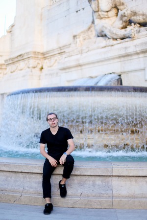 Young businessman sitting near Trevi fountain, wearing glasses and black clothes. Concept of youth, traveling and spending time in city.の写真素材