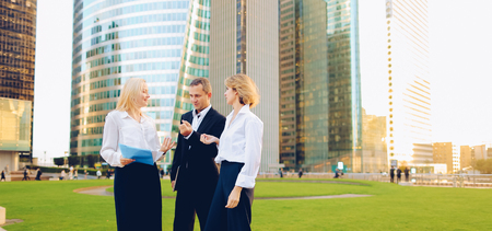 Beautiful businesswomen keeping blue document case talking with cheerful male boss outside. Concept of speaking with partners and joking about work. Pretty ladies dressed in white shirts smiling with man.の写真素材