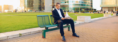 Businessperson reading newspaper and looking at watch outdoors in  . Concept of having break, resting and mass media. Prosperous man wears dark suit with white shirt.の写真素材