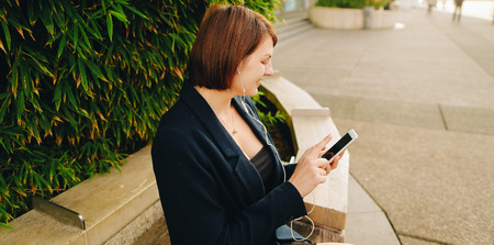 Businesswoman listening to music and chatting with smartphone and in ear phones outside. Concept of...