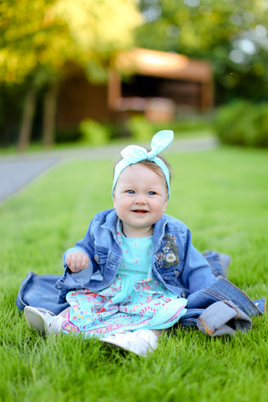 Little smiling female baby sitting on green grass and wearing jeans jacket. Concept of childhood and resting in garden, nature background.の写真素材