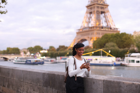 Happy business woman standing on bridge not far from Eiffel Tower and watching pictures of little daughter received from husband using mobile. Lady went to Paris for work. Young mother with beautiful hair styling wearing spacious backpack. Concept of business trip, international cooperation, innovative technologiesの写真素材