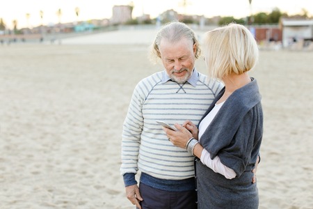 Elderly couple using smartphone on sand beach. Concept of pensioners and modern technology.