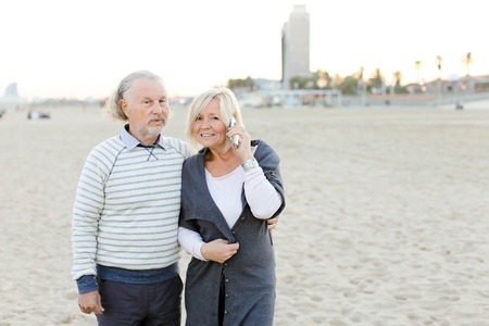 Senior husband walking on sand beach with blonde wife speaking by smartphone. Concept of health way of life and modern technology, pensioners.の写真素材