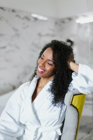Afro american smiling woman sitting in bathroom and wearing white bathrobe. Concept of relax and...