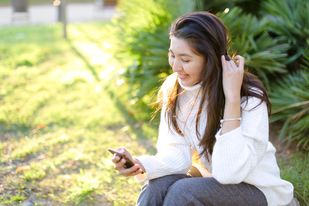 Japanese female student sitting in park and typing by smartphone. Concept of modern technology and social networks.の写真素材
