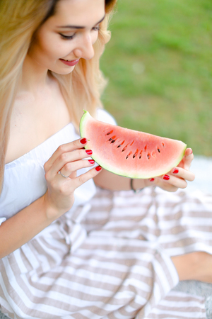 Young caucasian girl eating watermelon and wearing dress, grass in background. Concept of summer photo session, picnic.の写真素材