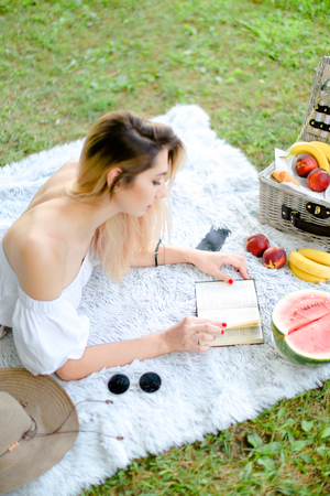 Young blonde woman reading book and lying on plaid near fruits and hat, grass in background. Concept of summer picnic and resting on weekends in open air.の写真素材