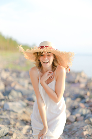 Young smiling woman in hat with red ribbon standing on shingle beach. Concept of summer vacations and resort.の写真素材