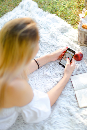 Young caucasian girl with red nails lying in park on plaid and enjoying social networks by smartphone. Concept of having free time, picnic and modern technology.の写真素材