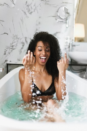 Young jocund afro american girl sitting in bathroom with marble tile, wearing black swimsuit....