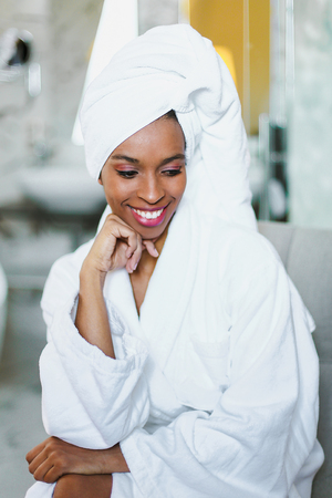 Young cute afro american woman sitting in bathroom and wearing white bathrobe. Concept of relax and personal care.の写真素材