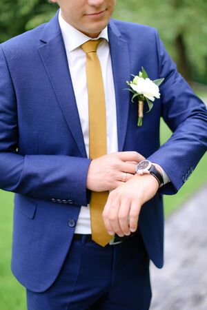 Young groom looking at watch and wearing blue jacket with flower. Concept of wedding photo session and waiting for bride.の写真素材