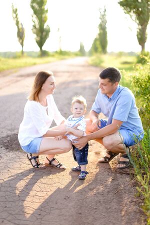 Happy young mother and father sitting on road with little baby, sunshine weather. Concept of parents and children, resting on nature.の写真素材