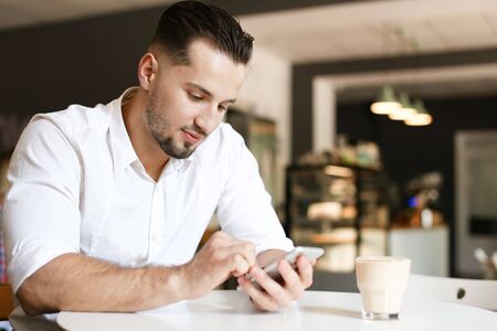 Young businessman chatting by smartphone at cafe, wearing white shirt. Concept of business and modern technology, free hotspot.の写真素材