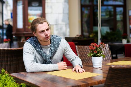 Young caucasian man sitting at street cafe near green plants, wearing grey sweater. Concept of resting in open air and waiting lunch.の写真素材