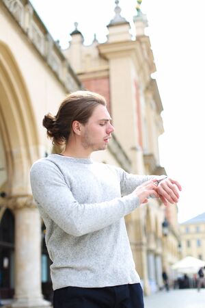 Young man looking at watch walking on square in Europe, wearing grey sweater. Concept of visiting Europe and tourist.の写真素材