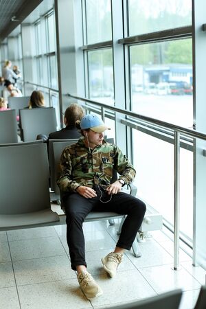 Caucasian male student sitting in waiting room at airport and listening to music with white earphones, wearing camouflage shirt and blue cap. Concept of traveling and cheap tickets, law cost flights.の写真素材