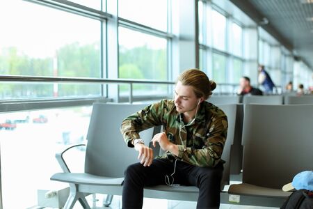 Man with long hair wearing camouflage shirt, sitting in waiting hall at airport near window, listening to music with white earphones and looking at watch. Concept of low cost flights and traveling.の写真素材