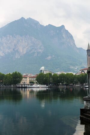 Calm morning coastline with buildings, mountain with clouds in background. Concept of pisturesque lake Como and traveling to italian countryside.の写真素材