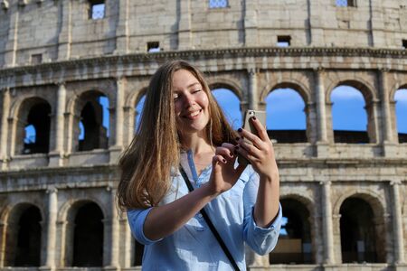 Young woman browsing by smartphone in Colosseum background in Rome, Italy. Concept of last minute tours to Italy and using Internet by modern gadget for social networks.の写真素材
